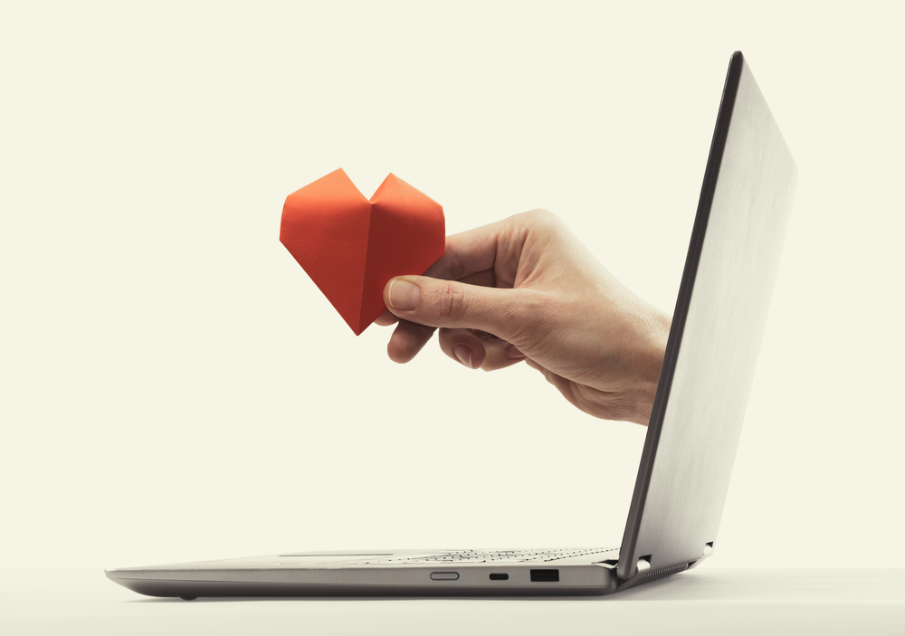 a hand holding a paper heart, reaching out of a laptop screen